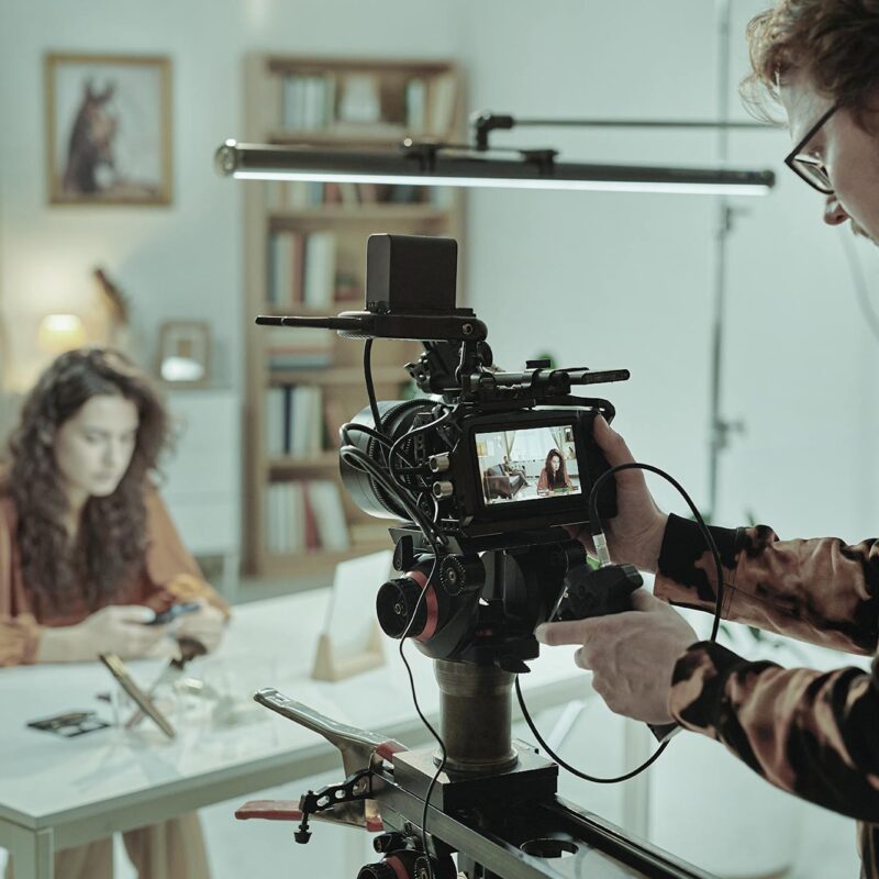 Behind the scenes af en videoproduktion til et E-Learning. Mand bag kamera optager kvinde ved bord.